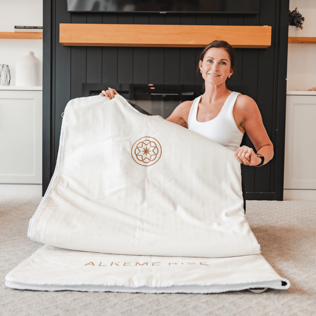 Founder of Alkemē Rise holding a white infrared sauna blanket in a calm home setting, representing real rituals and intentional wellness.