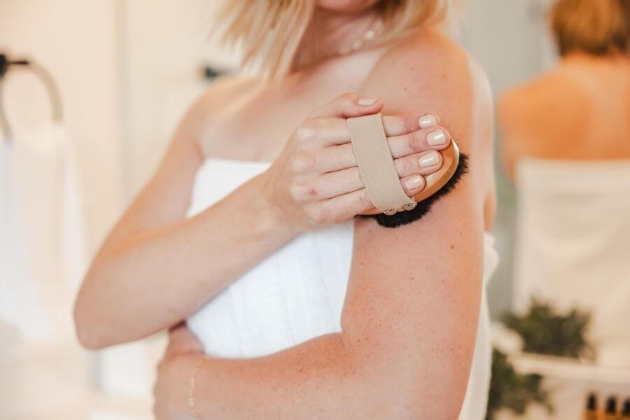 Close-up of a woman using the Alkemē Rise Copper Dry Brush on her upper arm before a shower, demonstrating a revitalizing self-care and natural detox pathway ritual.