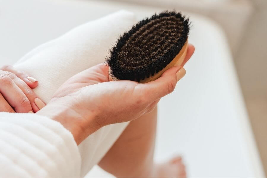 Close-up of hands holding the Alkemē Rise Ion Charged Copper Dry Brush over legs during a self-care ritual, highlighting the brush’s natural bristles and ergonomic shape.