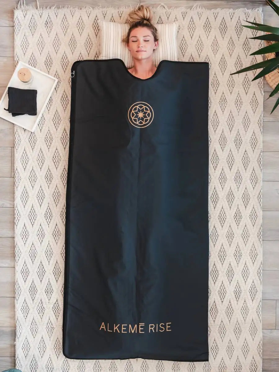 Woman lying peacefully in a white Alkeme Rise infrared sauna blanket with eyes closed, resting on a natural fiber mat with a digital controller visible beside her.