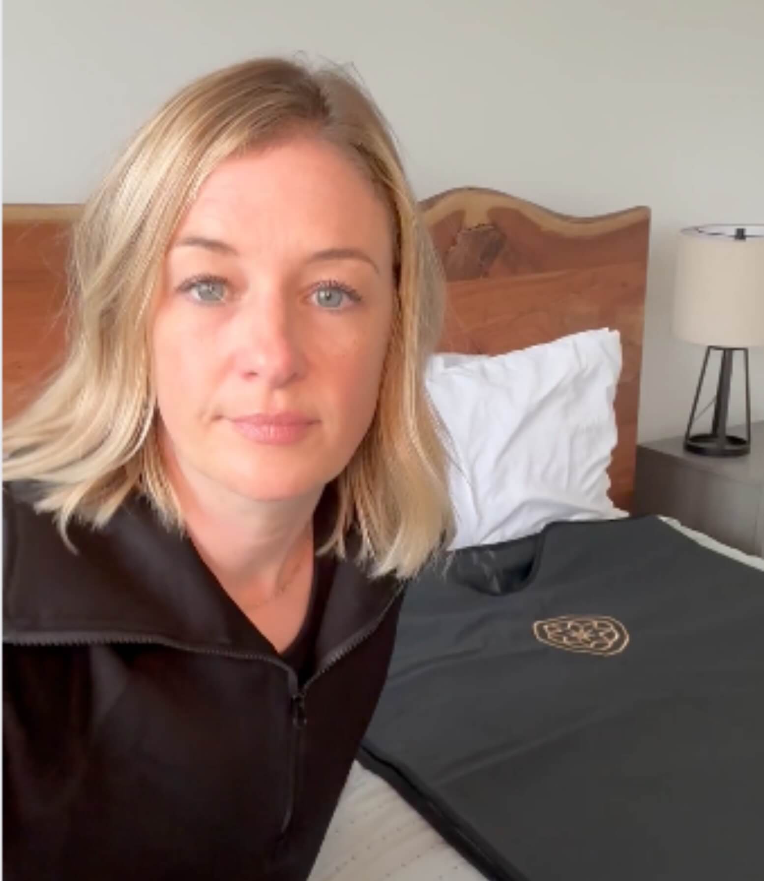 Blonde women sits at the edge of her bed with the Alkēme Rise sauna blanket draped over her shoulders, showing the elegant gold logo and ready-to-use setup.