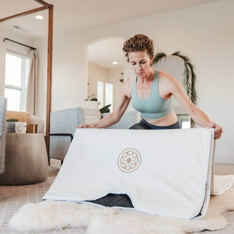 Woman setting up Alkeme Rise sauna blanket on a bedroom floor as part of pre-session ritual. Preparing to add her towel insert.