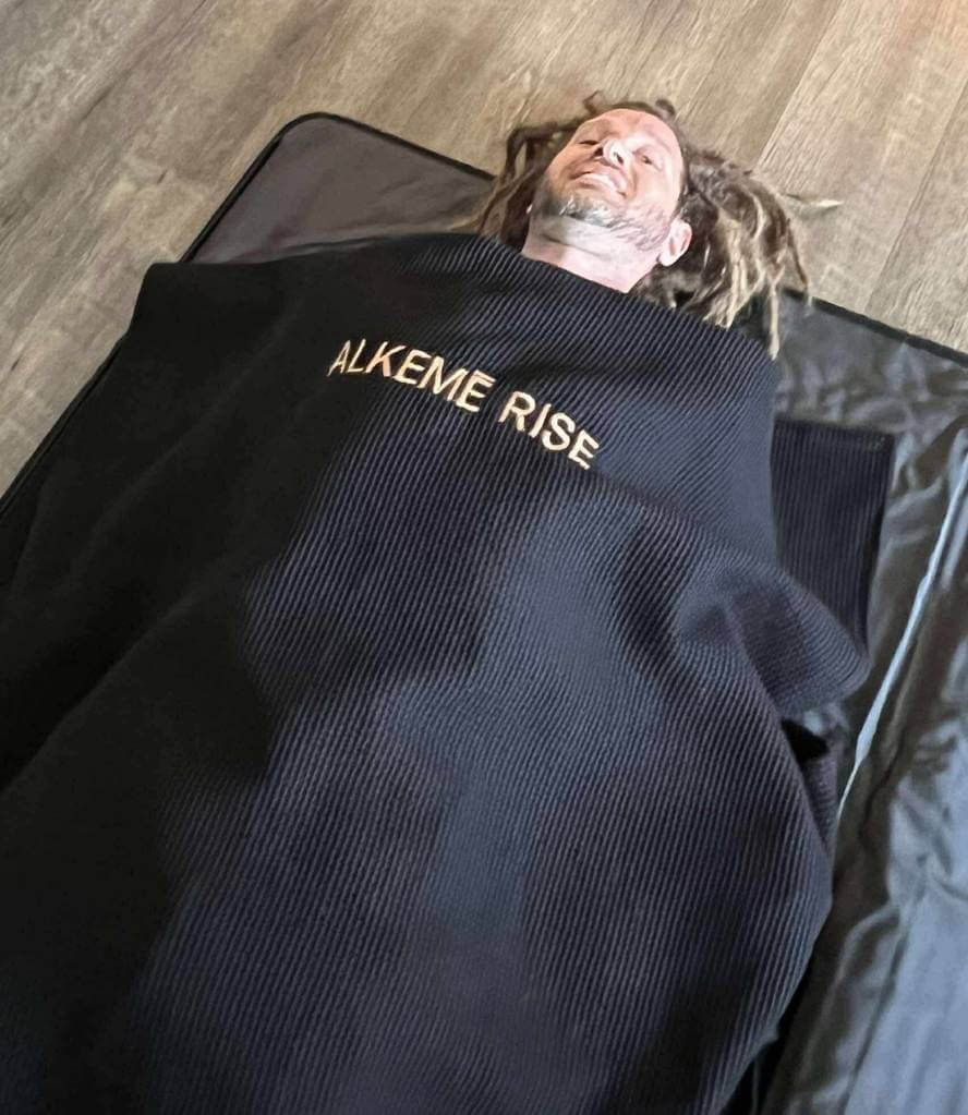 Man relaxes fully inside the Alkēme Rise infrared sauna blanket with a black towel insert placed on top. The setup highlights comfort and ease of use.