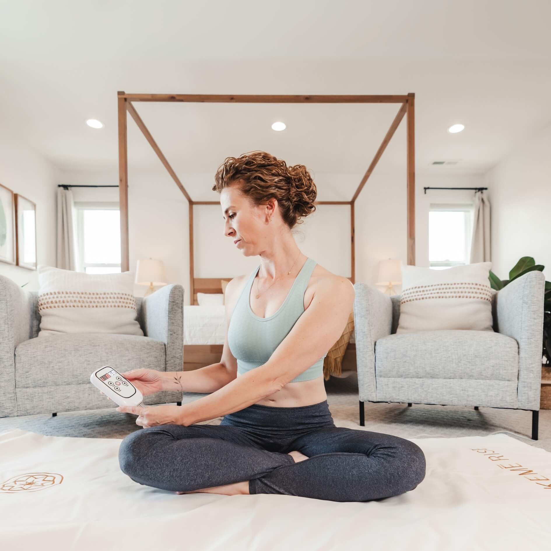 Woman sitting on the Alkēme Rise infrared sauna blanket remote, demonstrating ease of use and wellness from home.