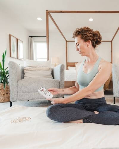 Woman seated on a bed using a white infrared sauna blanket with a remote, showing how simple and intuitive the product is to use at home.