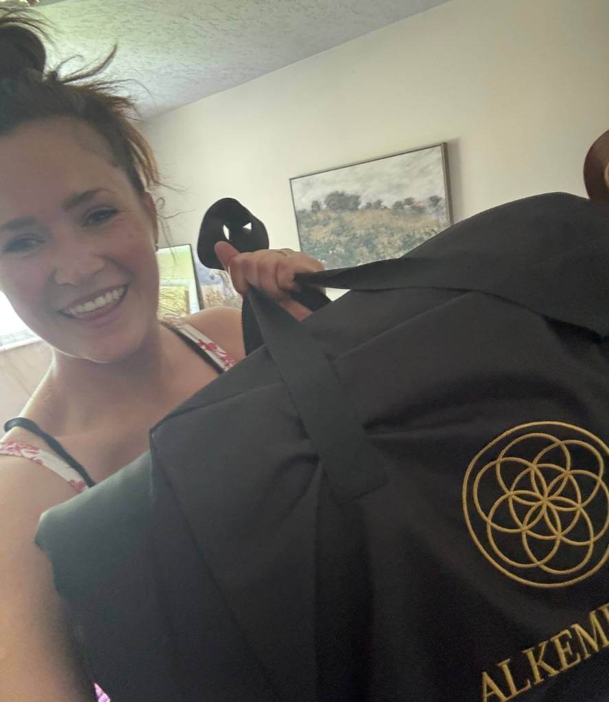 Women happily displays the Alkēme Rise infrared sauna blanket, showing the gold sacred geometry logo and folded blanket in a cozy, homey setting.