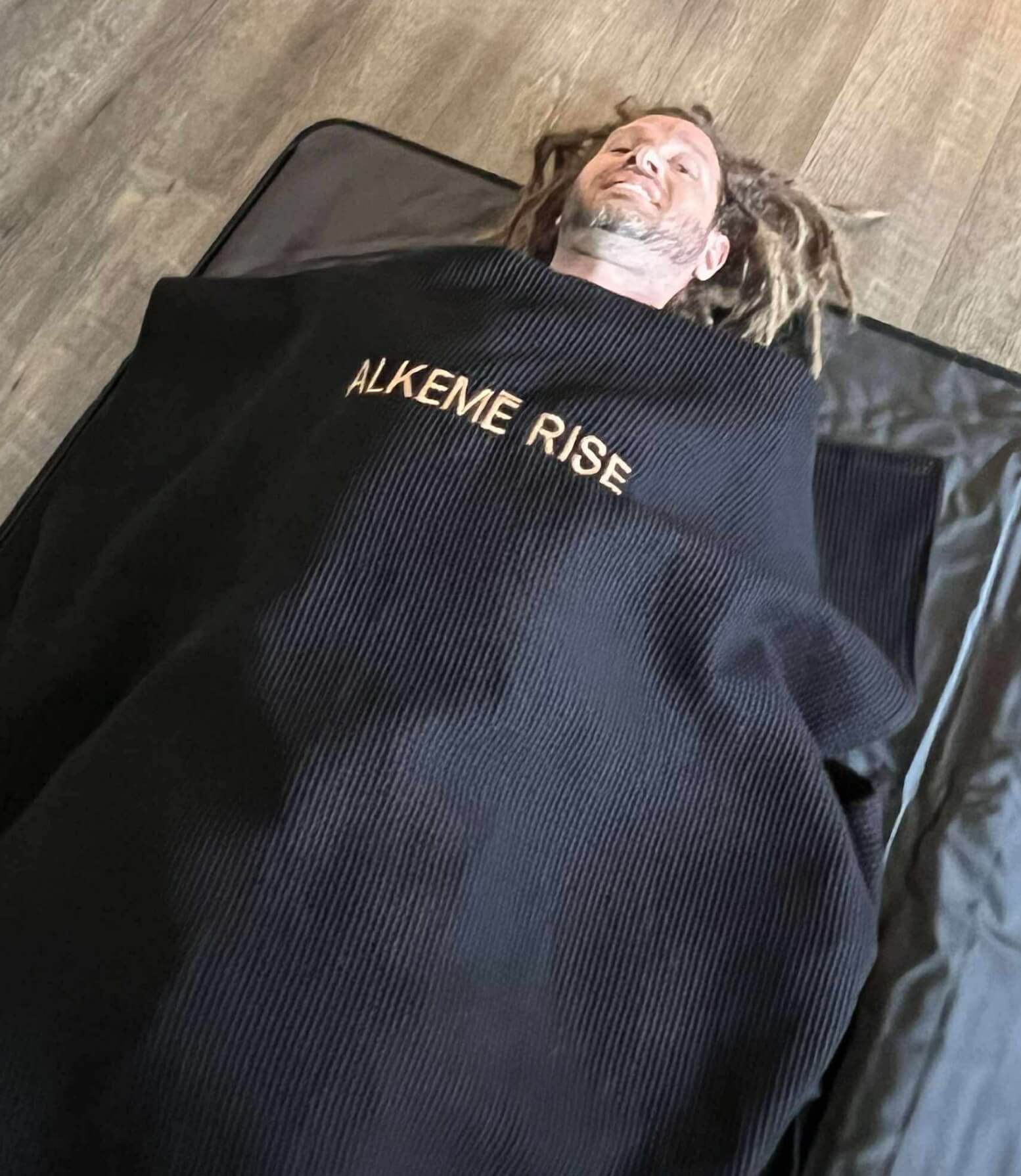 Man relaxes fully inside the Alkēme Rise infrared sauna blanket with a black towel insert placed on top. The setup highlights comfort and ease of use.