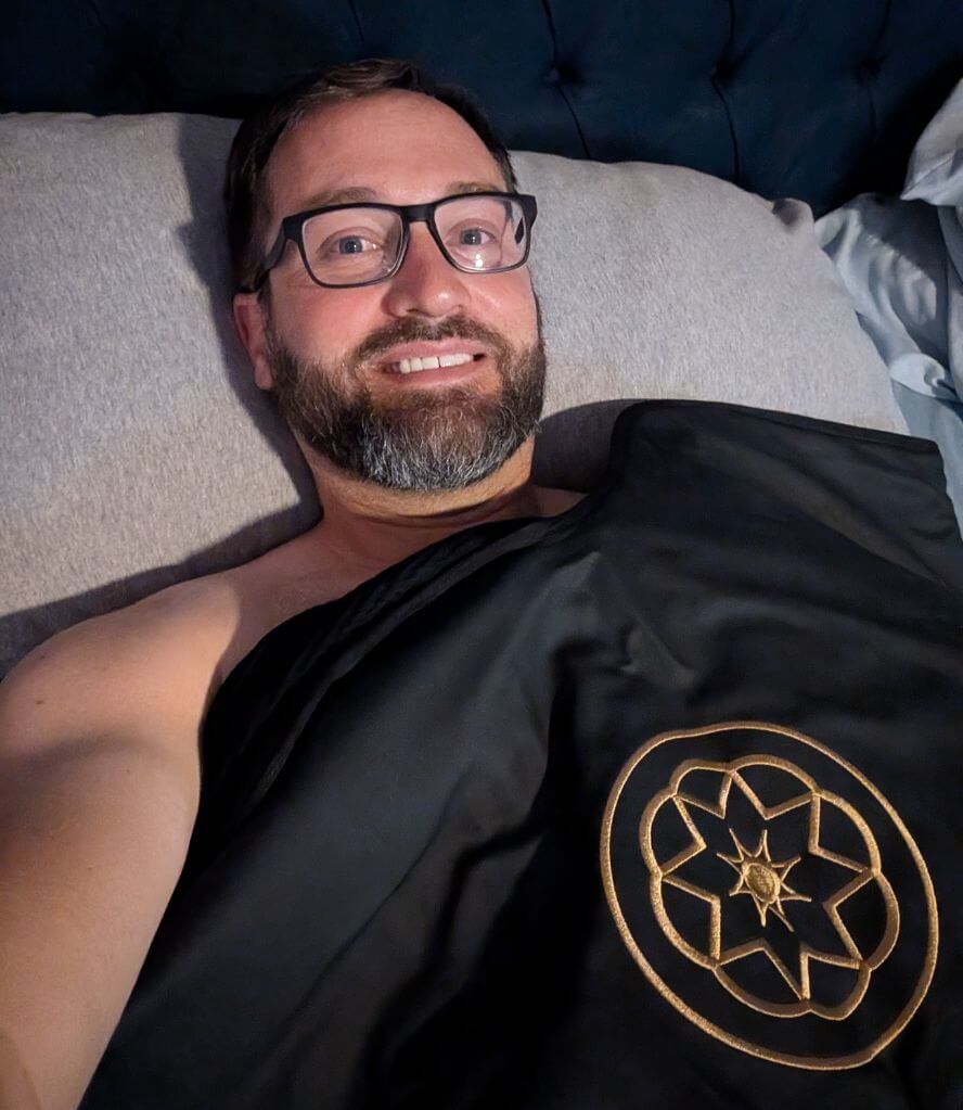 Man smiles while using the black Alkēme Rise infrared sauna blanket in bed. The gold sacred geometry logo is visible as he relaxes during his wellness ritual.