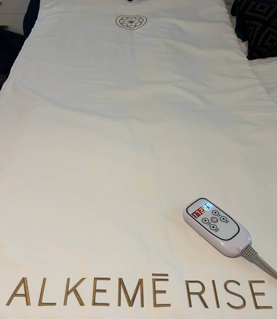 Full view of the Alkemē Rise Infrared Sauna Blanket laid out with remote, featuring the elegant logo and premium towel insert.
