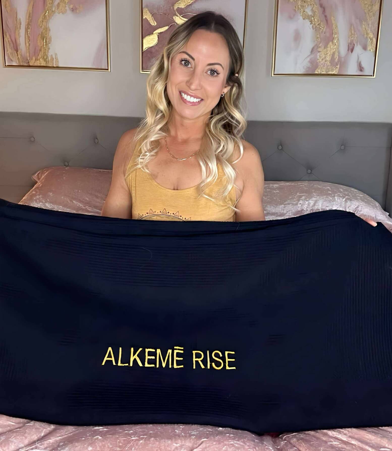 Blonde women sits on her bed holding up the black Alkēme Rise towel insert with gold embroidery. She smiles as she showcases the premium cotton layer used for sauna sessions.
