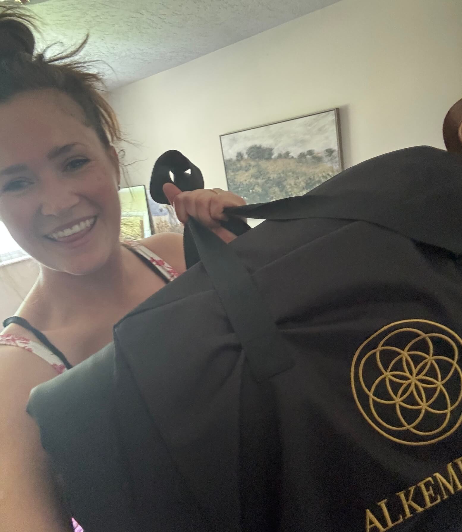 Women happily displays the Alkēme Rise infrared sauna blanket, showing the gold sacred geometry logo and folded blanket in a cozy, homey setting.