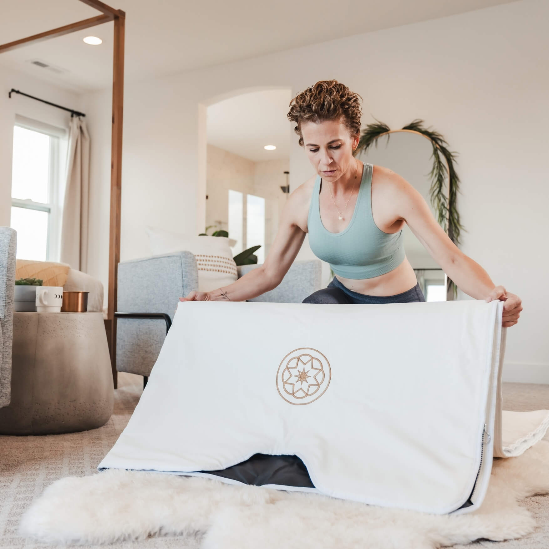 Woman setting up Alkeme Rise sauna blanket on a bedroom floor as part of pre-session ritual. Preparing to add her towel insert.