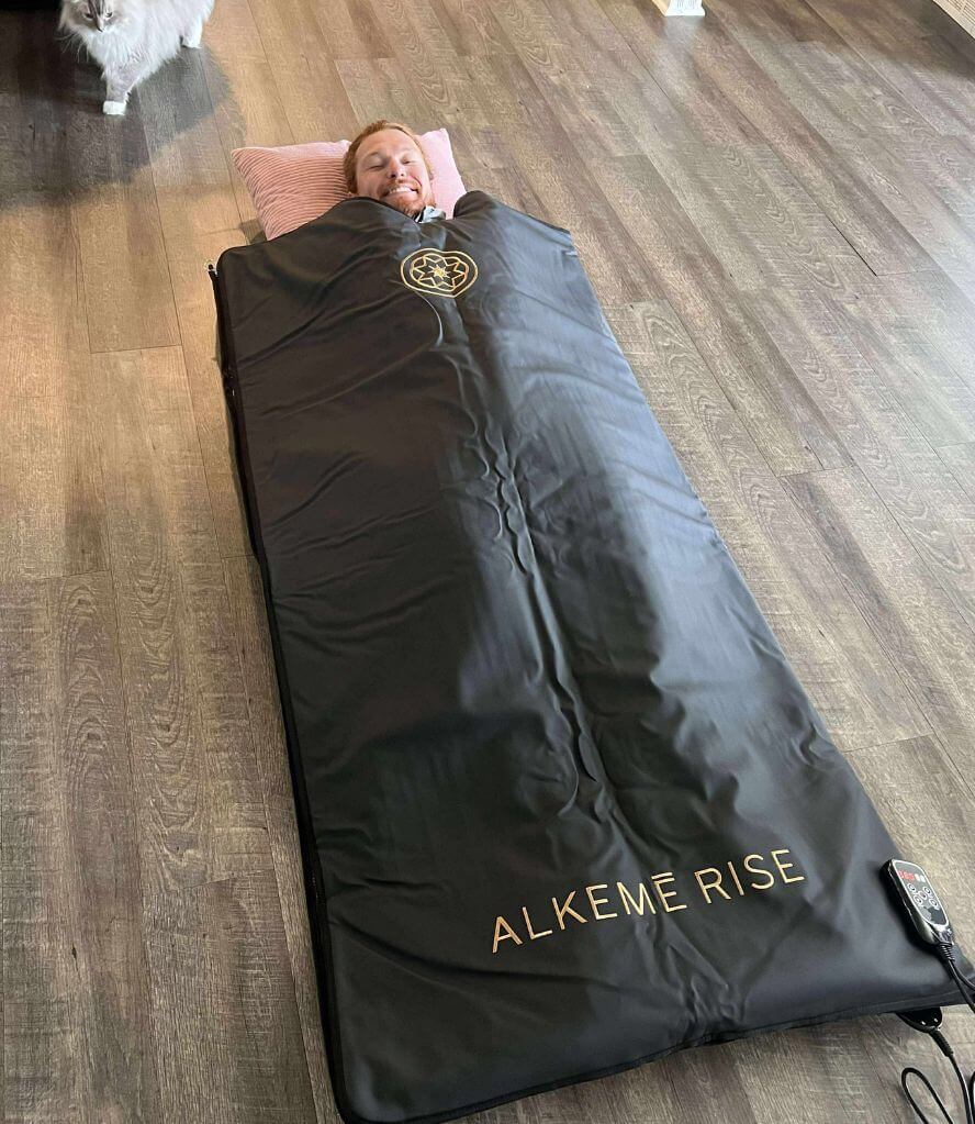 Man lies comfortably zipped into a black Alkēme Rise infrared sauna blanket on a wooden floor, showing the full-body fit and cozy design during an at-home wellness session.