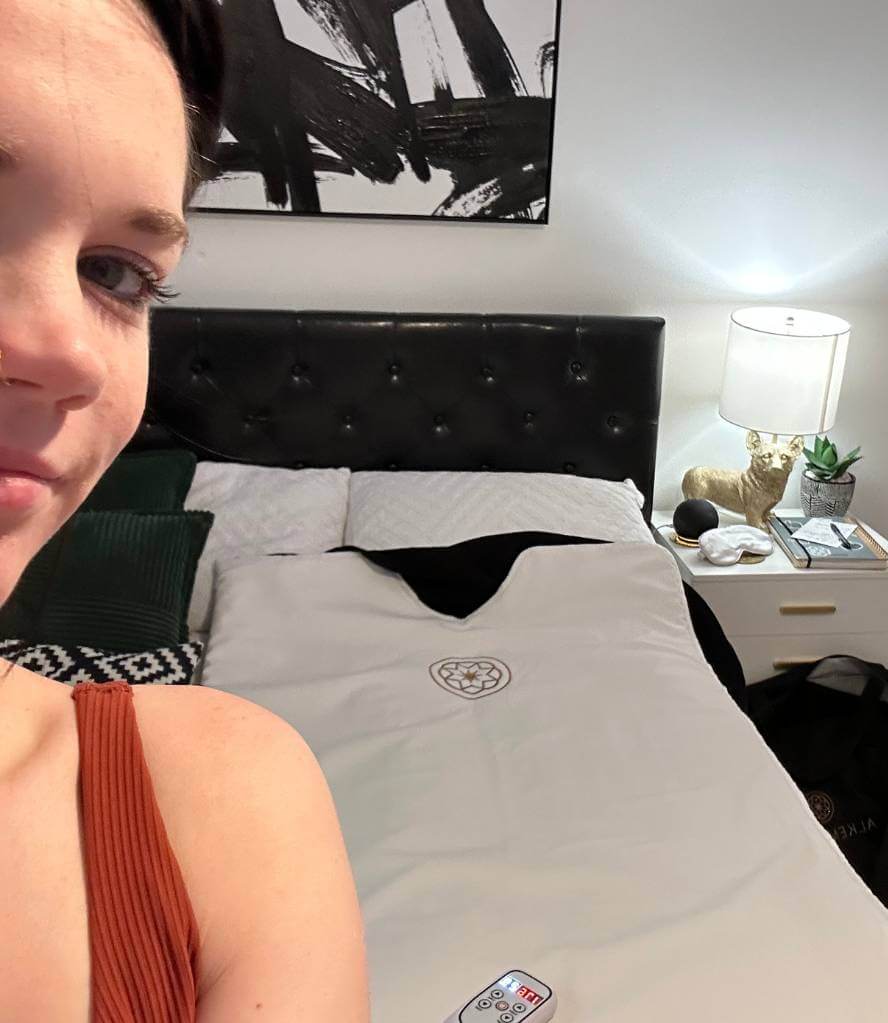 Real customer enjoying a wellness ritual with the Alkemē Rise Infrared Sauna Blanket set up in a cozy bedroom setting.