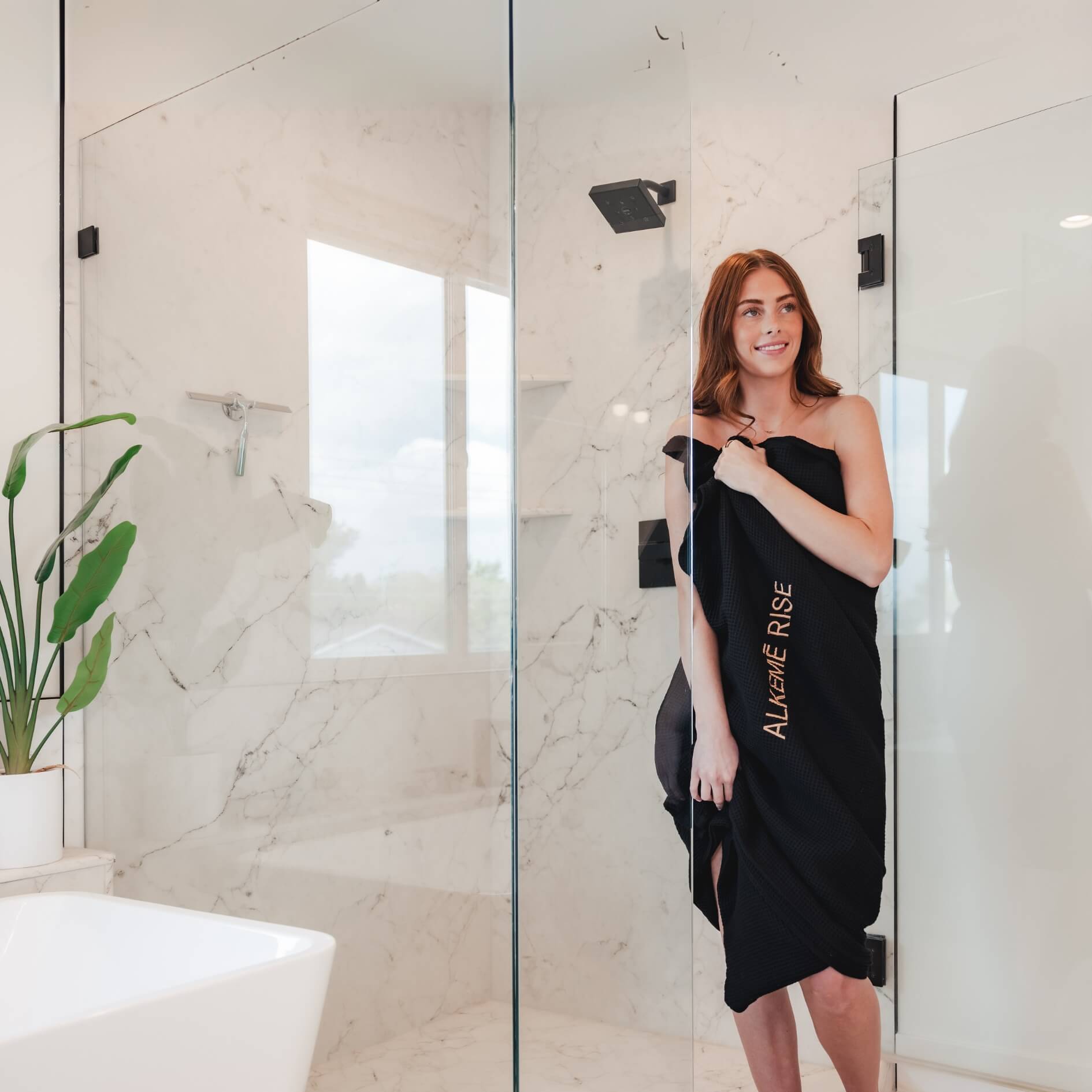 Woman standing in bathroom wrapped in Alkēme Rise towel insert after a sauna session, representing premium comfort and thoughtful design.